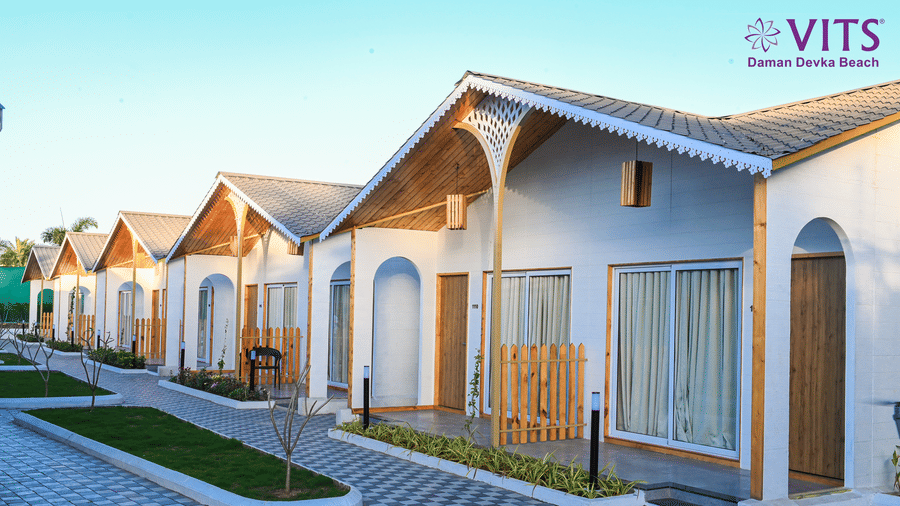 A view of the cottages' facade with cobbled pathway, a small lawn in between the pathway and wooden gates on the entrance to the cottage at VITS Daman Devka Beach.