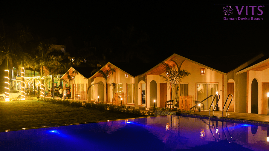 A well-lit resort facade at night, with a large swimming pool in front and warm yellow lights wrapped around palm trees for decoration | VITS Daman Devka Beach
