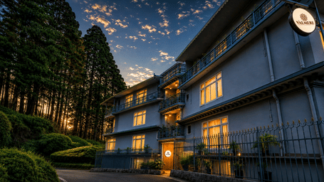 A multi-storey hotel building at dusk, illuminated windows, a circular signboard, and surrounding pine trees at Valmere Lungta Boutique Hotel & Spa, Darjeeling.