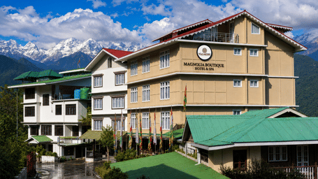 Daytime exterior of Valmere Magnolia Boutique Hotel & Spa, Pelling with its signboard, green roofs, prayer flags, and snow-capped Himalayan peaks behind.