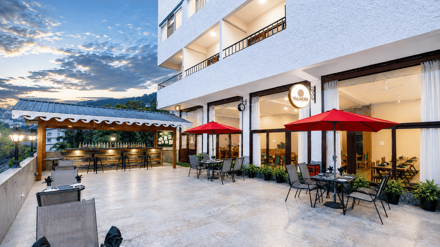 Exterior of Valmere Himalayan Hotel & Spa, Gangtok at dusk showing the building facade, an outdoor dining terrace with red umbrellas, and a covered bar area.
