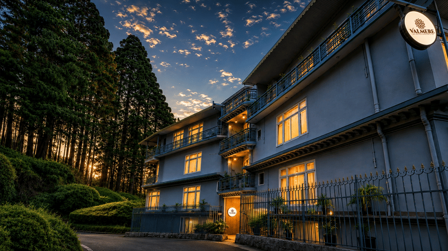 A multi-storey hotel building at dusk, illuminated windows, a circular signboard, and surrounding pine trees at Valmere Lungta Boutique Hotel & Spa, Darjeeling.