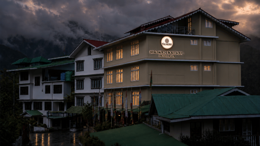 Twilight exterior of Valmere Magnolia Boutique Hotel & Spa, Pelling with illuminated windows and signage against a dramatic cloudy mountain sky.