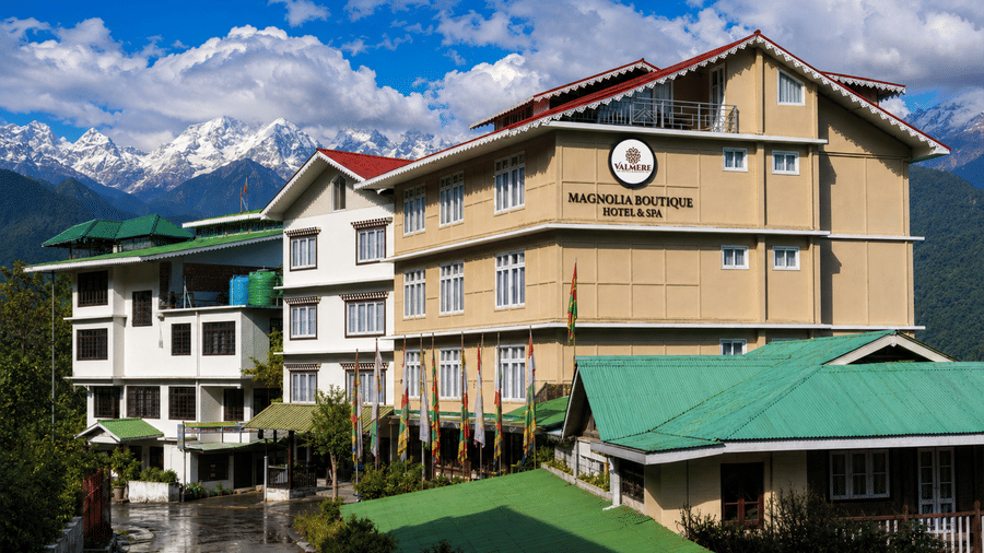 Daytime exterior of Valmere Magnolia Boutique Hotel & Spa, Pelling with its signboard, green roofs, prayer flags, and snow-capped Himalayan peaks behind.