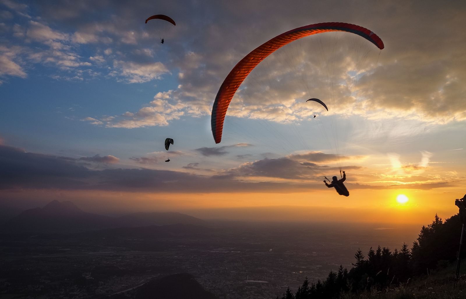 Image of a  para-glider soaring through the sky at sunset, with a breathtaking view of mountains and forest.