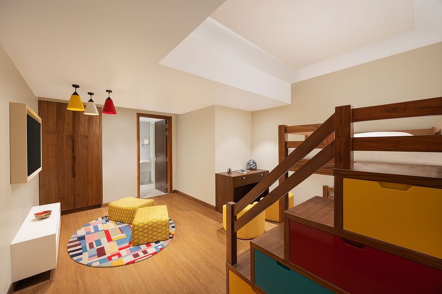 A vibrant children's bedroom featuring a wooden bunk bed with colourful drawers, a small wooden desk and yellow chair, a colourful rug with two yellow patterned poufs, wooden wardrobes, a wall-mounted television, and colourful pendant lights, with an open 