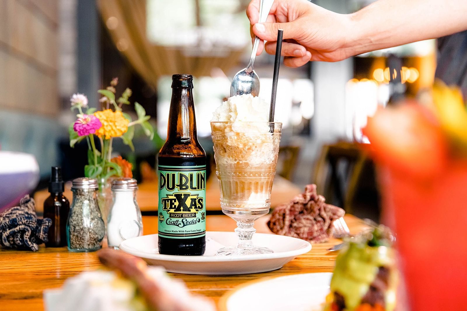 A hand holding a spoon digging inside a glass beside a bottle, placed in a a  plate, and small flowers on the table.- The Grand Hotel & Ballroom, McKinney