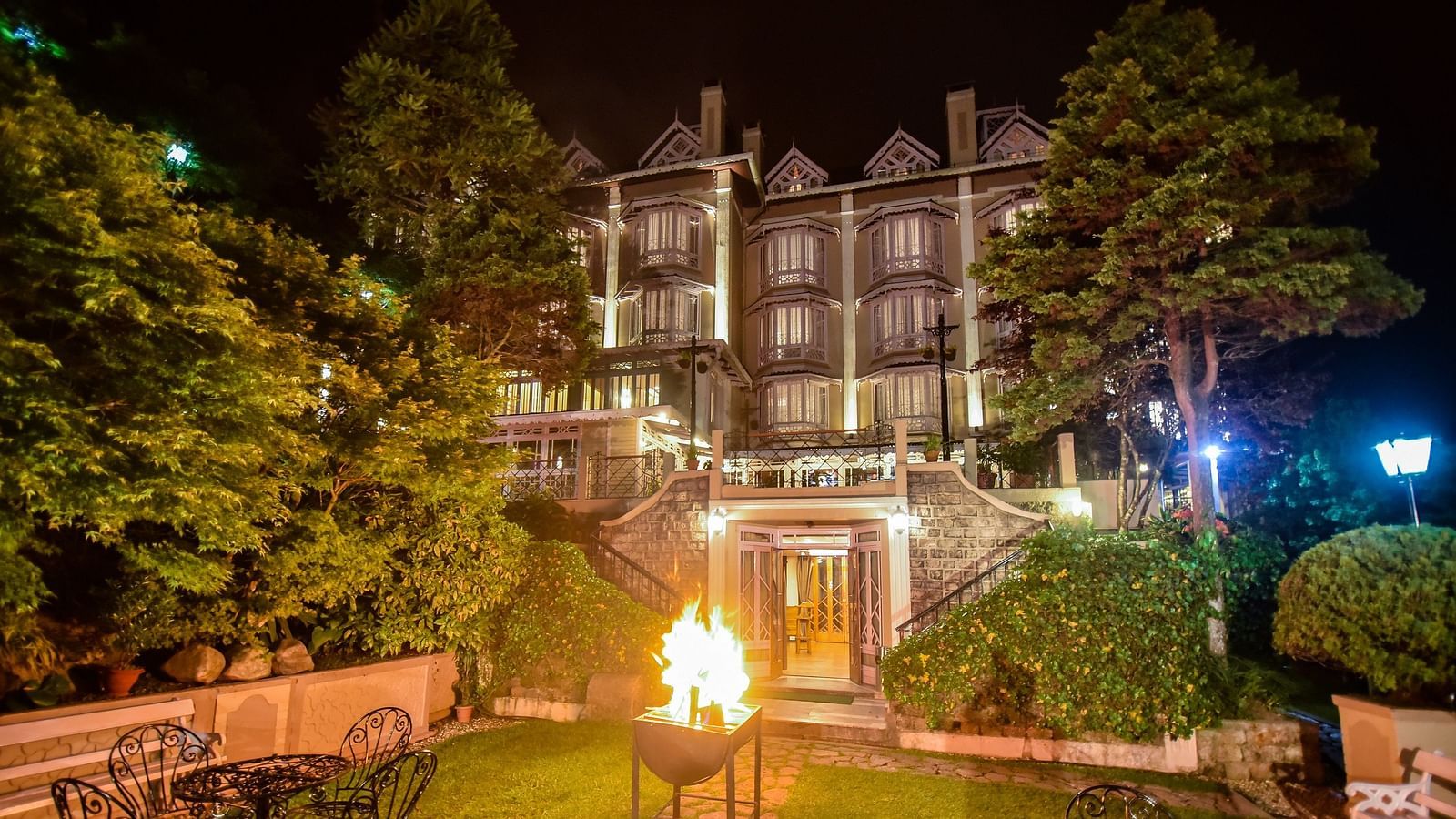 Night view of Cedar Inn, Darjeeling with a cozy outdoor fire pit, garden seating, and the warmly lit heritage building surrounded by lush greenery.