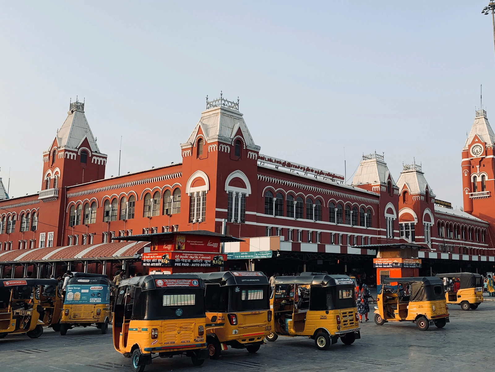 The iconic red Gothic-style facade of Chennai Central Railway Station, featuring its grand clock tower and several yellow auto-rickshaws parked in the foreground.