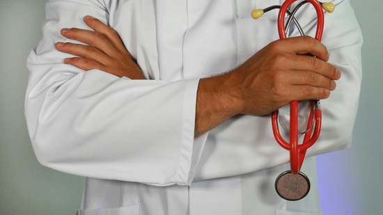 A medical professional in a lab coat stands with arms crossed, holding a stethoscope in one hand.