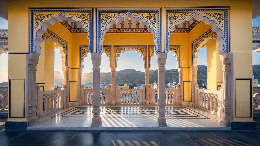 a balcony at the rooftop with yellow walls and intricate paintings on the pillars with blue colour with the sun rays brightening up the area - Brij Castle, Jaipur