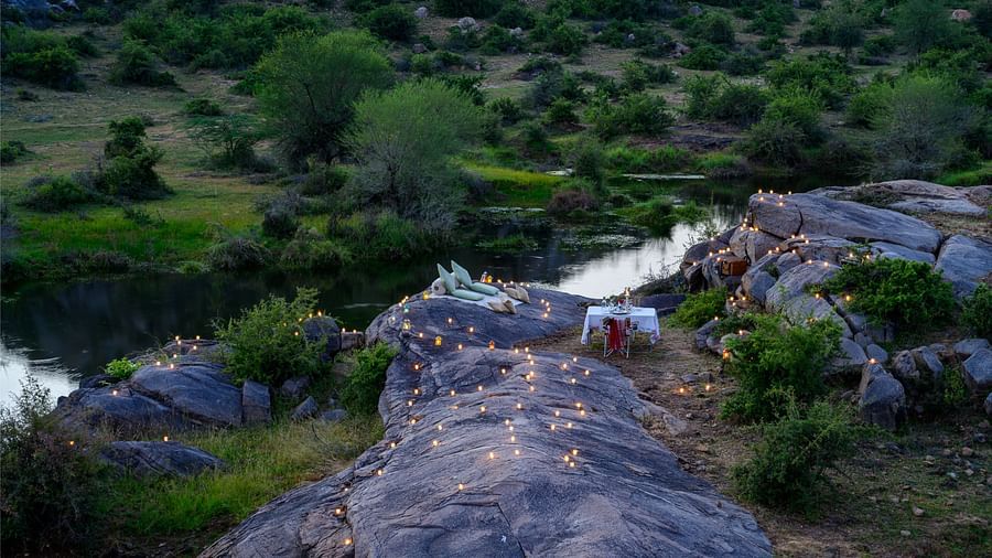 An image of an outdoor dining arrangement near a lake over viewing the mountains with bright lighting and greenery around - Utsav Camp Sariska