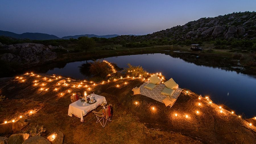 An image of an outdoor dining arrangement near a lake over viewing the mountains with bright lighting and greenery around - Utsav Camp Sariska