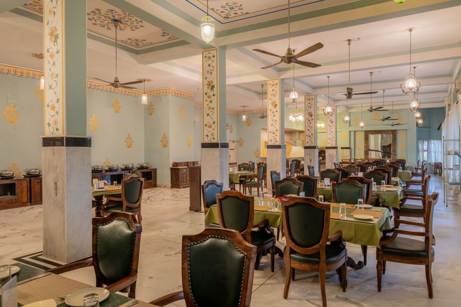 Fateh Mahal Restaurant  at Umaid Palace featuring sets of tables and chairs, ceiling fans, hanging lights, and a cabinet with food utensils at one corner.