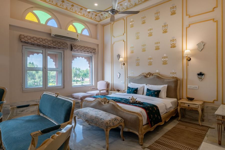 Maharaja Suite at Umaid Palace featuring a lavish bedroom with king size bedside table and lamps on either side. The room also have few soft sofa chairs an, a table, and a window near AC.