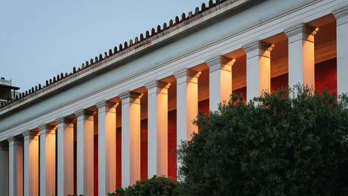 The exterior of a large, classical building showing a row of columns illuminated with a warm light.