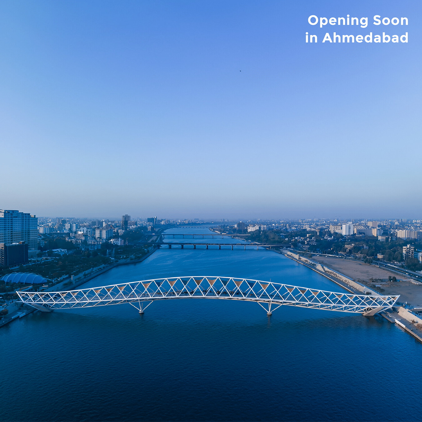 An image of a bridge above the crystal blue river under the clear sky as the text says 'Opening Soon in Ahmedabad'