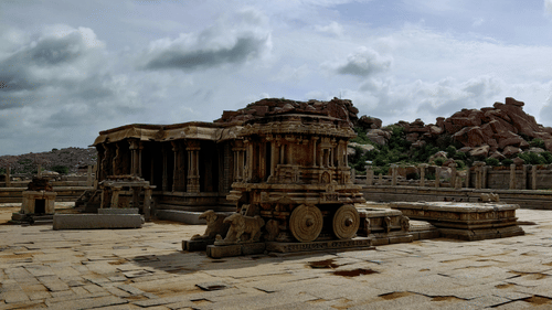 Weathered stone temple ruins in Hampi, one of the popular travelling places in Karnataka, stand across a vast courtyard, reflecting exquisite ancient craftsmanship amid boulder-strewn landscapes and cloudy skies overhead.