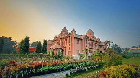 Pink heritage building in Agartala with twin domes, arched windows and a flowering nursery garden in the foreground at golden hour.