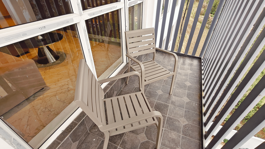 2 grey metal chairs are placed on a dark-tiled balcony beside a glass sliding door at Bliss Casa Mila By Bergamont Hotels, Kodaikanal.