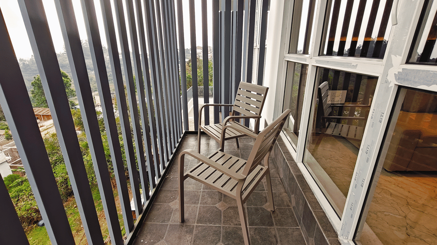 2 chairs sit on a balcony with a view of green trees through vertical grey metal railings at Bliss Casa Mila By Bergamont Hotels, Kodaikanal.