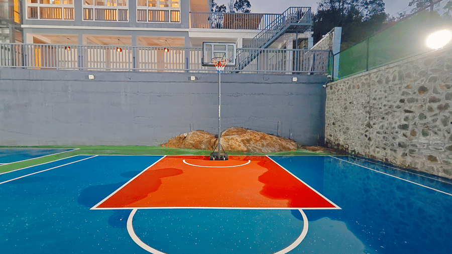 A blue court features a bright red rectangular key area under a single basketball hoop at Bliss Casa Mila By Bergamont Hotels, Kodaikanal.