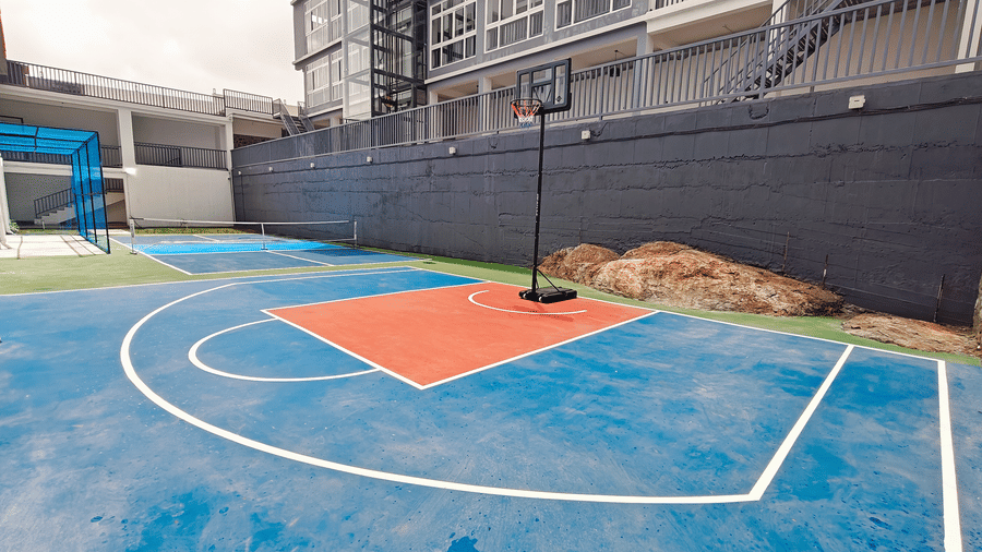 A basketball hoop stands on a blue and orange court next to a grey stone wall at Bliss Casa Mila By Bergamont Hotels, Kodaikanal.