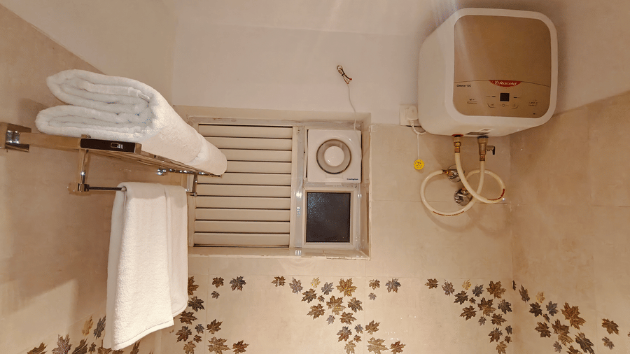 2 white towels rest on a metal rack near a small window and a white water heater on a tiled wall at Bliss Casa Mila By Bergamont Hotels, Kodaikanal.