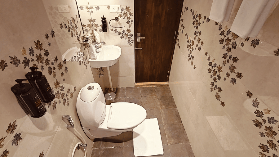 A white toilet and washbasin in a bathroom with leaf-patterned tiles and a dark wood door at Bliss Casa Mila By Bergamont Hotels, Kodaikanal.