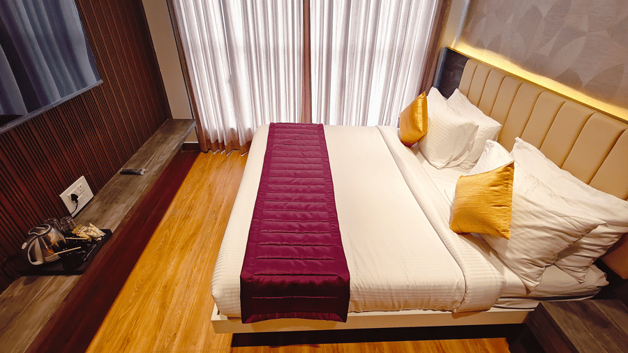 A double bed with a maroon runner is positioned next to a window with closed white curtains at Bliss Casa Mila By Bergamont Hotels, Kodaikanal.