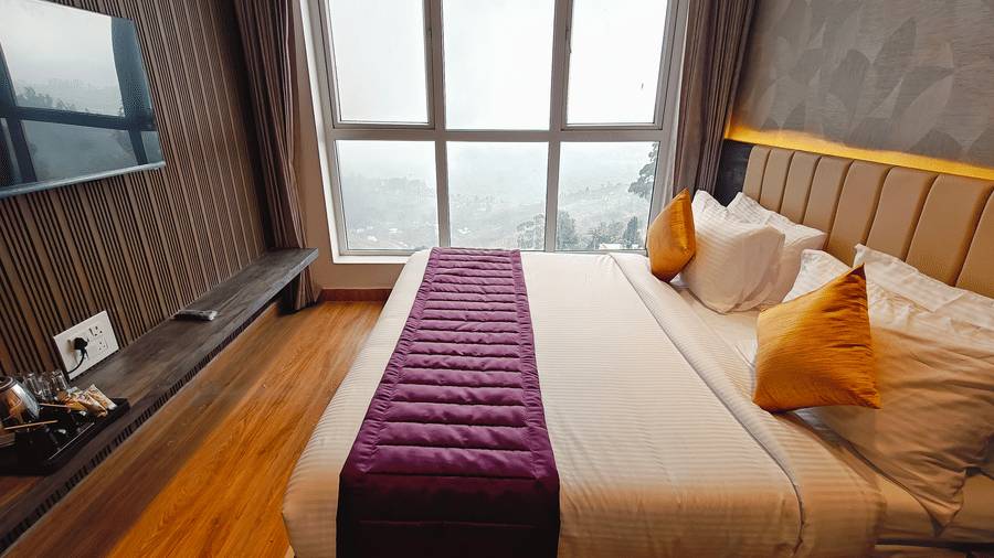 A double bed with yellow cushions and a purple runner faces a large window overlooking a misty landscape at Bliss Casa Mila By Bergamont Hotels, Kodaikanal.