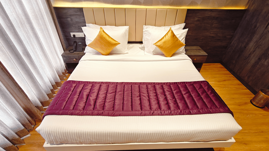 A white double bed with 2 yellow cushions and a purple runner is positioned between 2 bedside tables at Bliss Casa Mila By Bergamont Hotels, Kodaikanal.