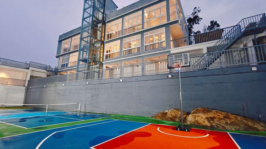 The illuminated 3-storey glass-fronted hotel building and basketball hoop at dusk at Bliss Casa Mila By Bergamont Hotels, Kodaikanal.
