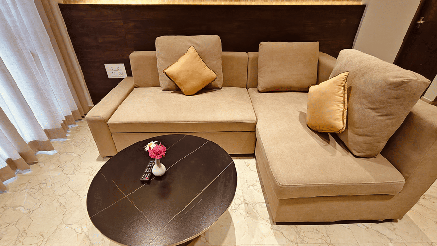 A beige L-shaped sofa with yellow cushions sits behind a round dark coffee table with a pink flower at Bliss Casa Mila By Bergamont Hotels, Kodaikanal.