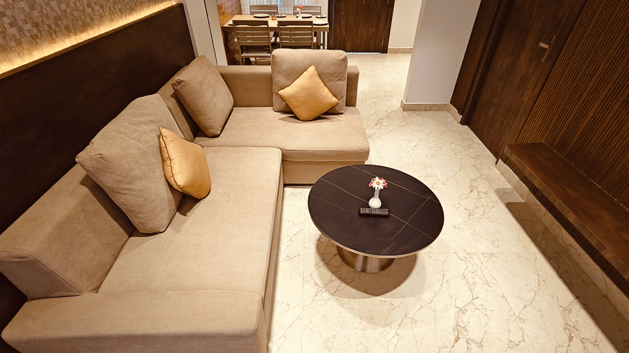 A beige L-shaped sofa with gold cushions facing a small round black coffee table on a marble floor at Bliss Casa Mila By Bergamont Hotels, Kodaikanal.