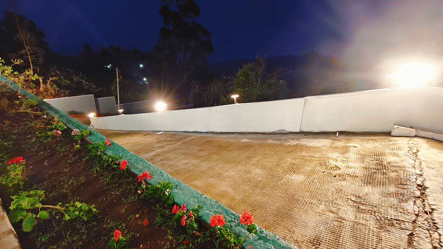 A white wall with 3 glowing lamps and a flower garden containing red flowers at night at Bliss Casa Mila By Bergamont Hotels, Kodaikanal.