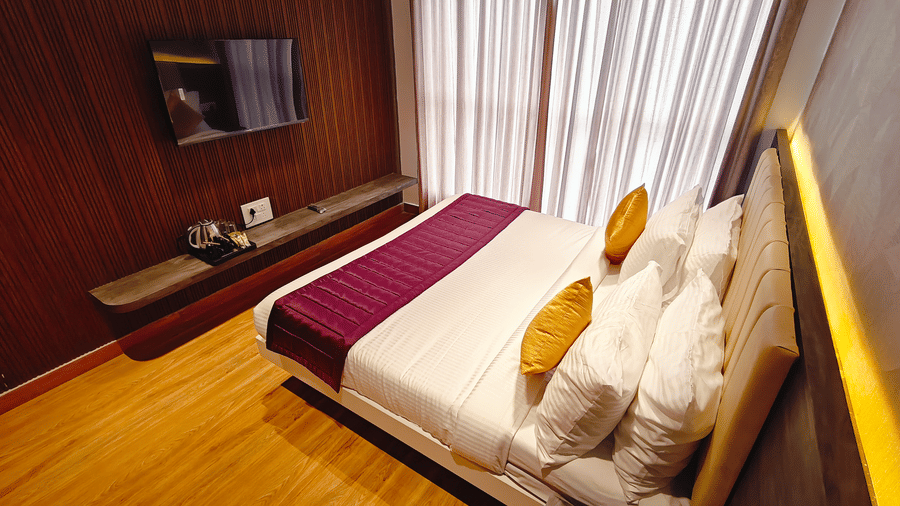 A high-angle view shows a double bed, a wall-mounted television, and a long wooden shelf at Bliss Casa Mila By Bergamont Hotels, Kodaikanal.