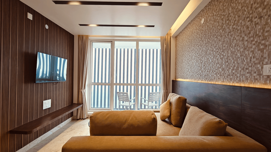 A television is mounted on a wooden wall opposite a beige sofa with large windows leading to a balcony at Bliss Casa Mila By Bergamont Hotels, Kodaikanal.