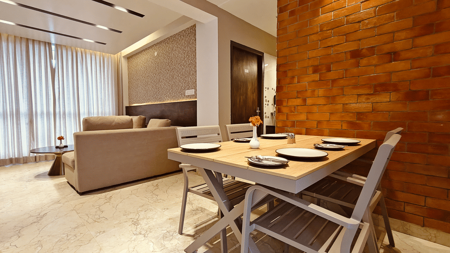 A dining table for 4 is situated in an open-plan suite next to a beige sofa and a red brick wall at Bliss Casa Mila By Bergamont Hotels, Kodaikanal.