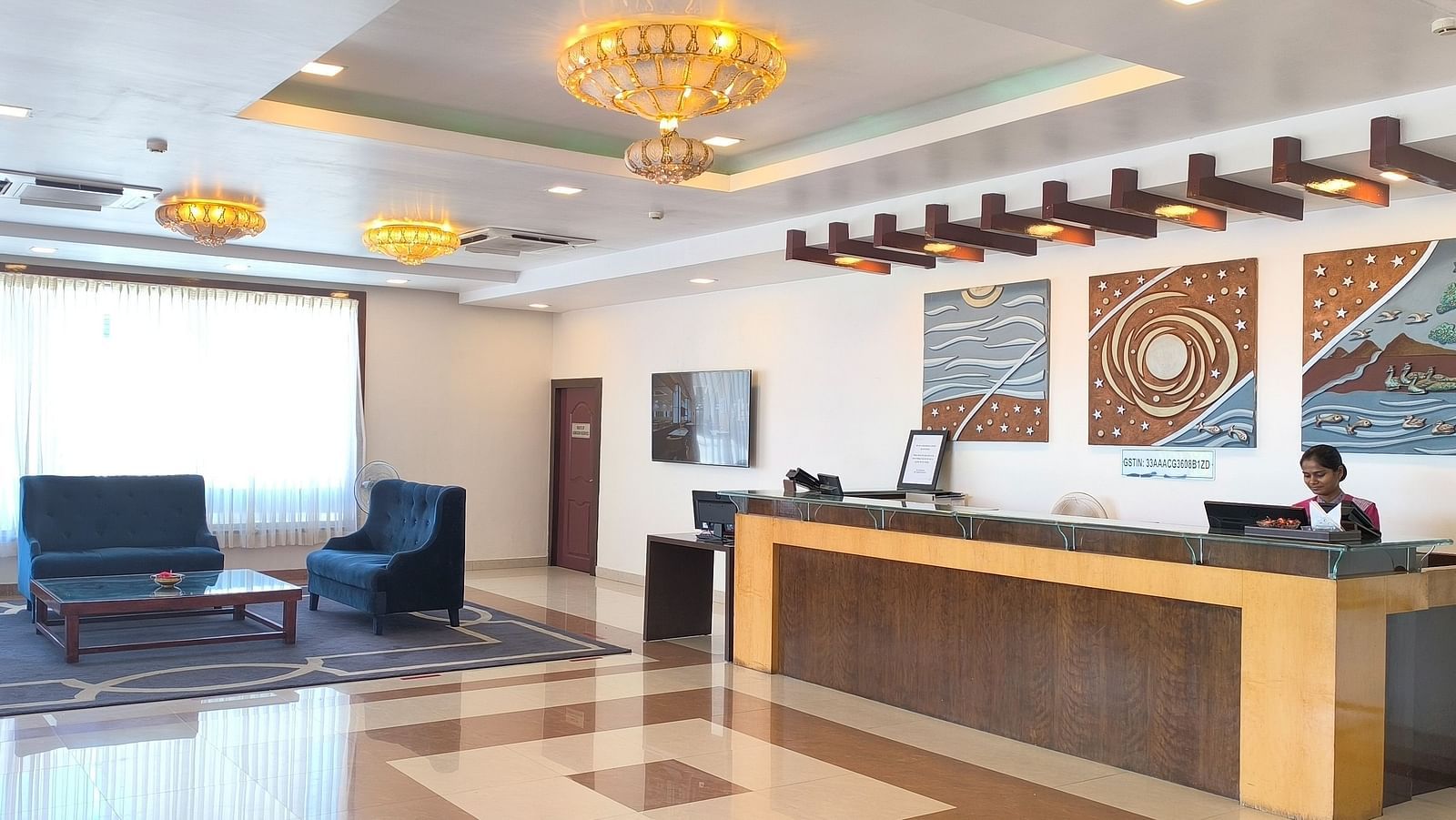An image of a hotel lobby featuring a reception desk with a receptionist standing behind it, and blue couches positioned under a warmly lit chandelier on the side - Regency Tuticorin by GRT Hotels