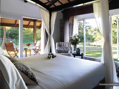 A bright, airy bedroom at The Residence, The Serai Kabini, with a canopy bed and sheer curtains, large windows opening to lush greenery, an armchair, and a covered patio with wooden chairs overlooking the garden for a relaxing stay in Kabini.