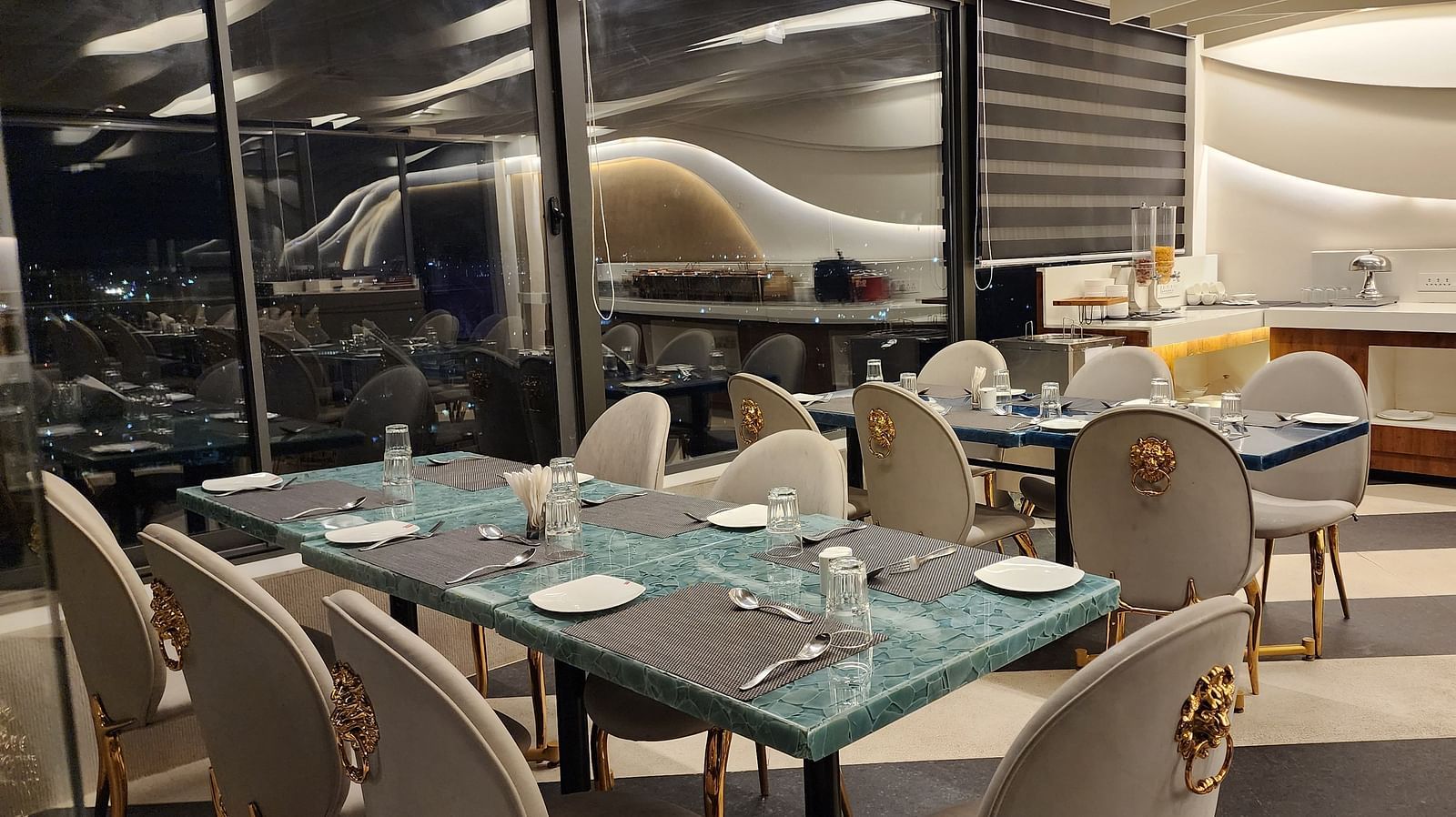 An elegant dining area with white and gold chairs and set tables, located beside large windows offering a view outside.