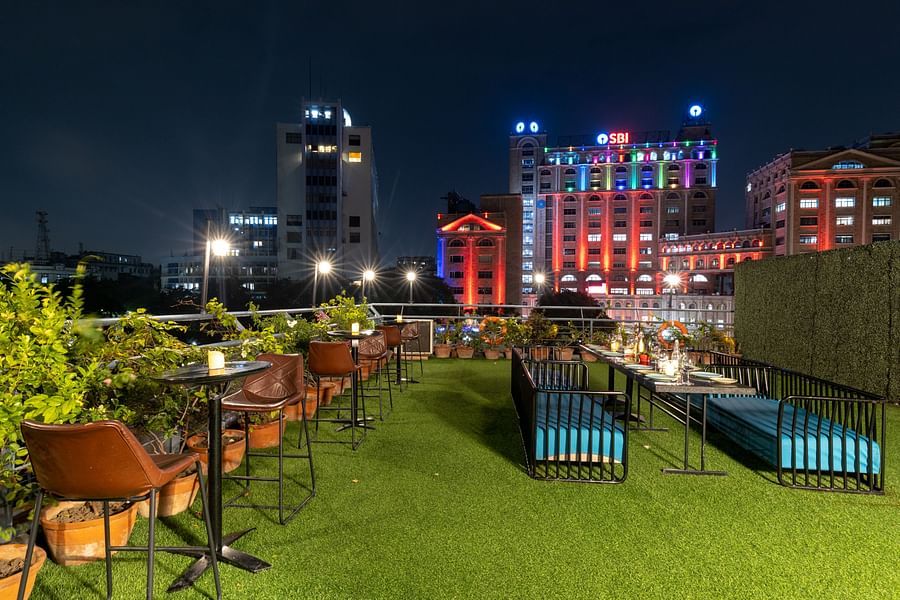 A nighttime view of a rooftop dining area with a green grass floor and a table set for a meal, city lights in the distance.