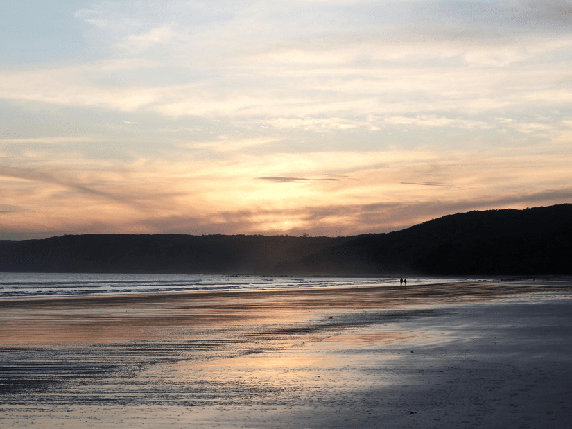 A beach while the sun is setting with the orange sky and other rocks at the back.