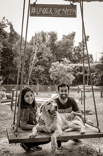 Two people sitting on a the ground with their pet dog on a swing under a sign reading Nature's Nook at Karma Chalets.