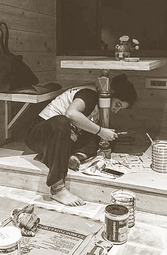 A person painting a table stand while sitting on the floor at Karma Chalets.