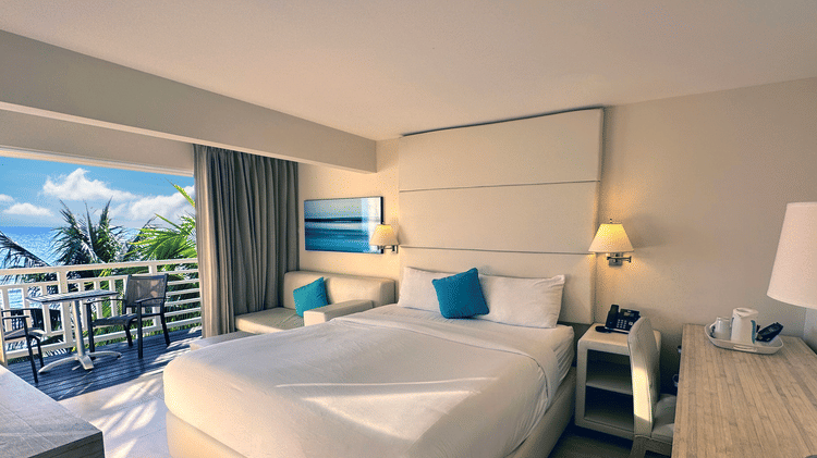 A neatly set room at The Soco Hotel featuring a large white bed with blue pillows, a lit lamp, and a balcony view of the ocean.