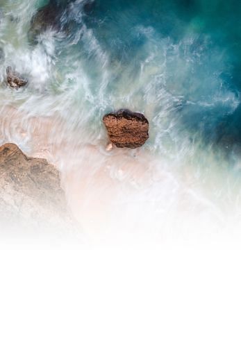 An aerial view of a coastal beach scene with waves crashing around a solitary large rock