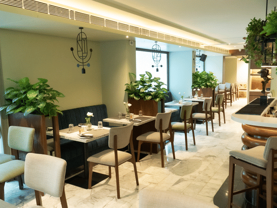A serene dining area at the Koi Garden of Hotel Bawa International in Vile Parle featuring neatly arranged tables, upholstered chairs, indoor plants, and soft ambient lighting creating a calm and elegant setting.