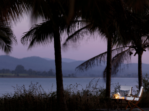 Two chairs and a table kept near the lakeside with coconut trees next to the chairs and faraway hills in view during twilight hour at The Serai Kabini - the best resort in Kabini.